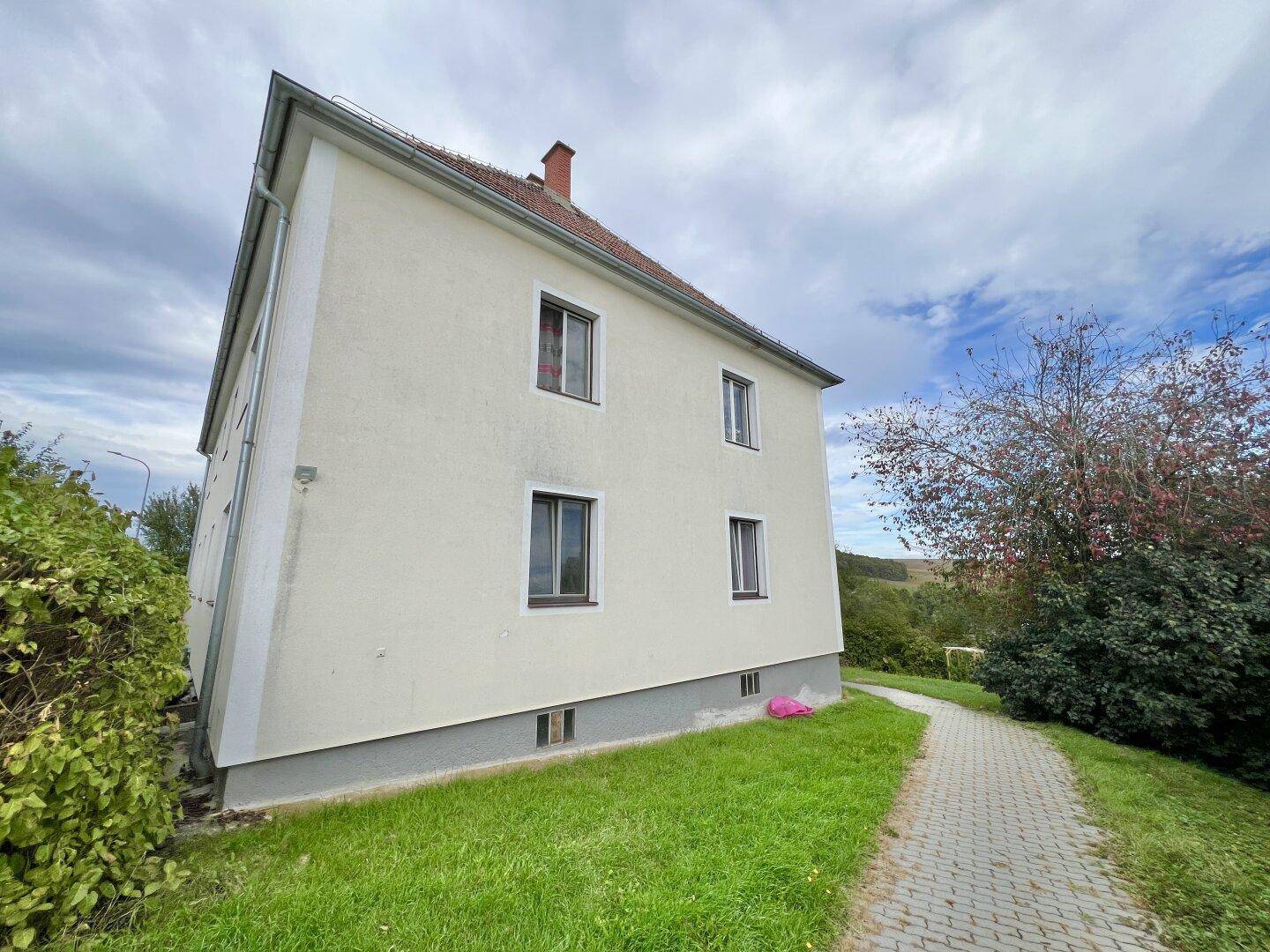 Seitenansicht des Hauses mit heller Fassade, Ziegeldach und einem gepflasterten Weg im Grünen.