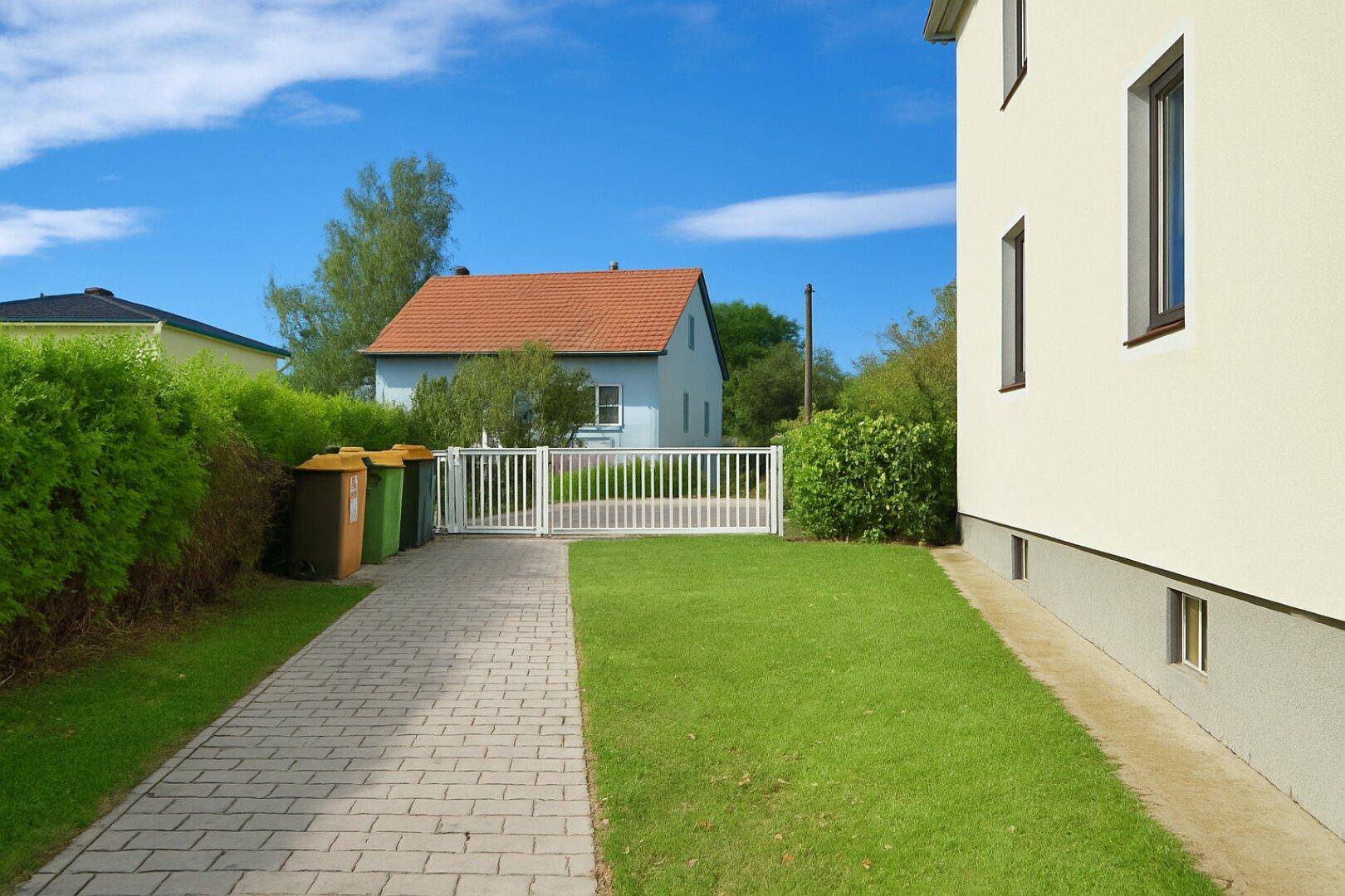 Einfahrt zum Grundstück mit weißem Tor, gepflastertem Weg und gepflegter Rasenfläche.