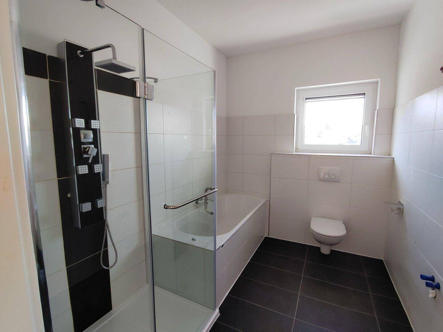 Modernes Badezimmer mit Duschkabine, Badewanne, WC und Fenster, ausgestattet mit dunklen Bodenfliesen.