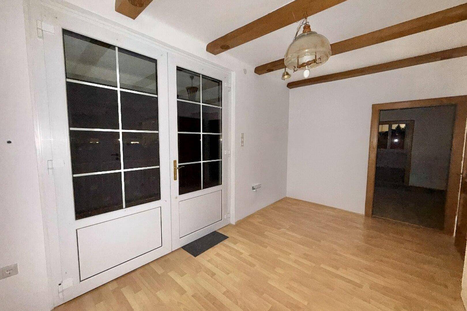 Heller Raum mit Holzbalkendecke und Zugang zu einem Außenbereich durch große Fenstertüren.