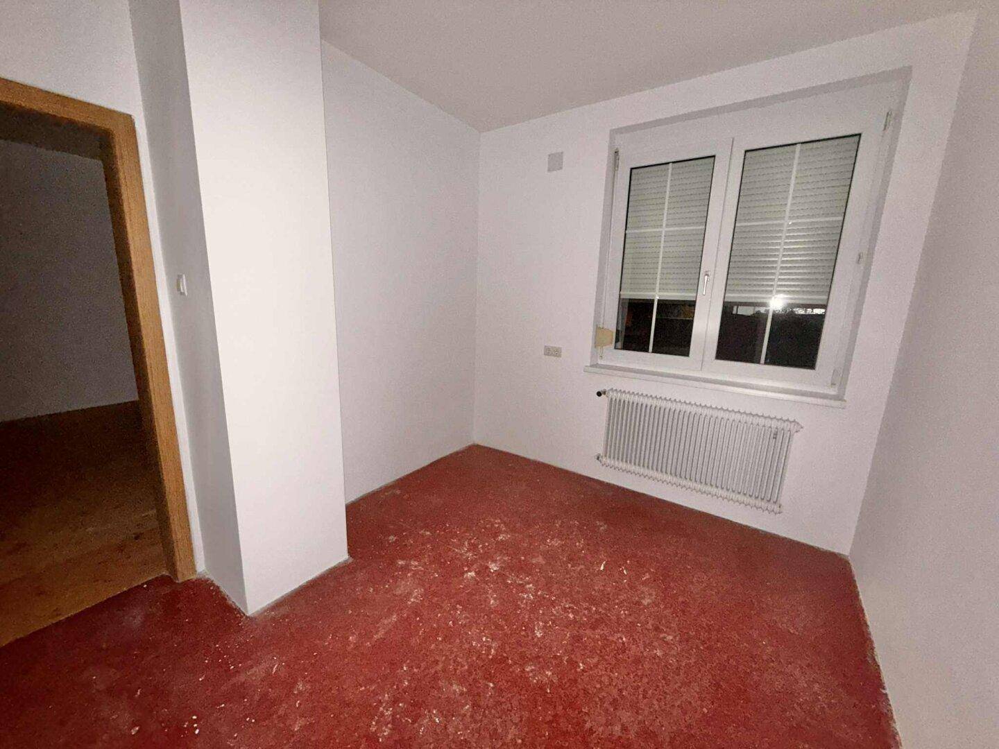 Kleiner Raum mit rotem Bodenbelag, weiß gestrichenen Wänden und Fenster mit Heizkörper.