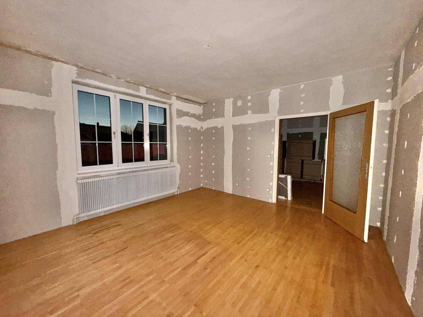 Renovierungsbedürftiger Raum mit Holzboden, vorbereiteten Wänden und Fenster.