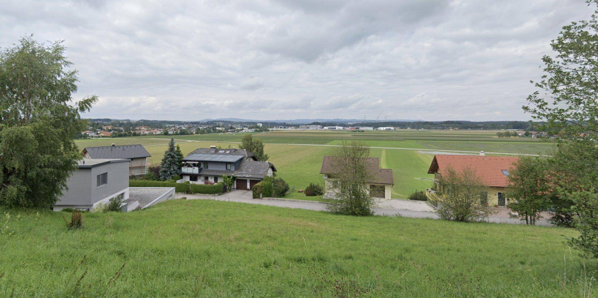 Blick auf eine ländliche Umgebung mit Wohnhäusern, weiten Feldern und einem Dorf in der Ferne unter bewölktem Himmel.