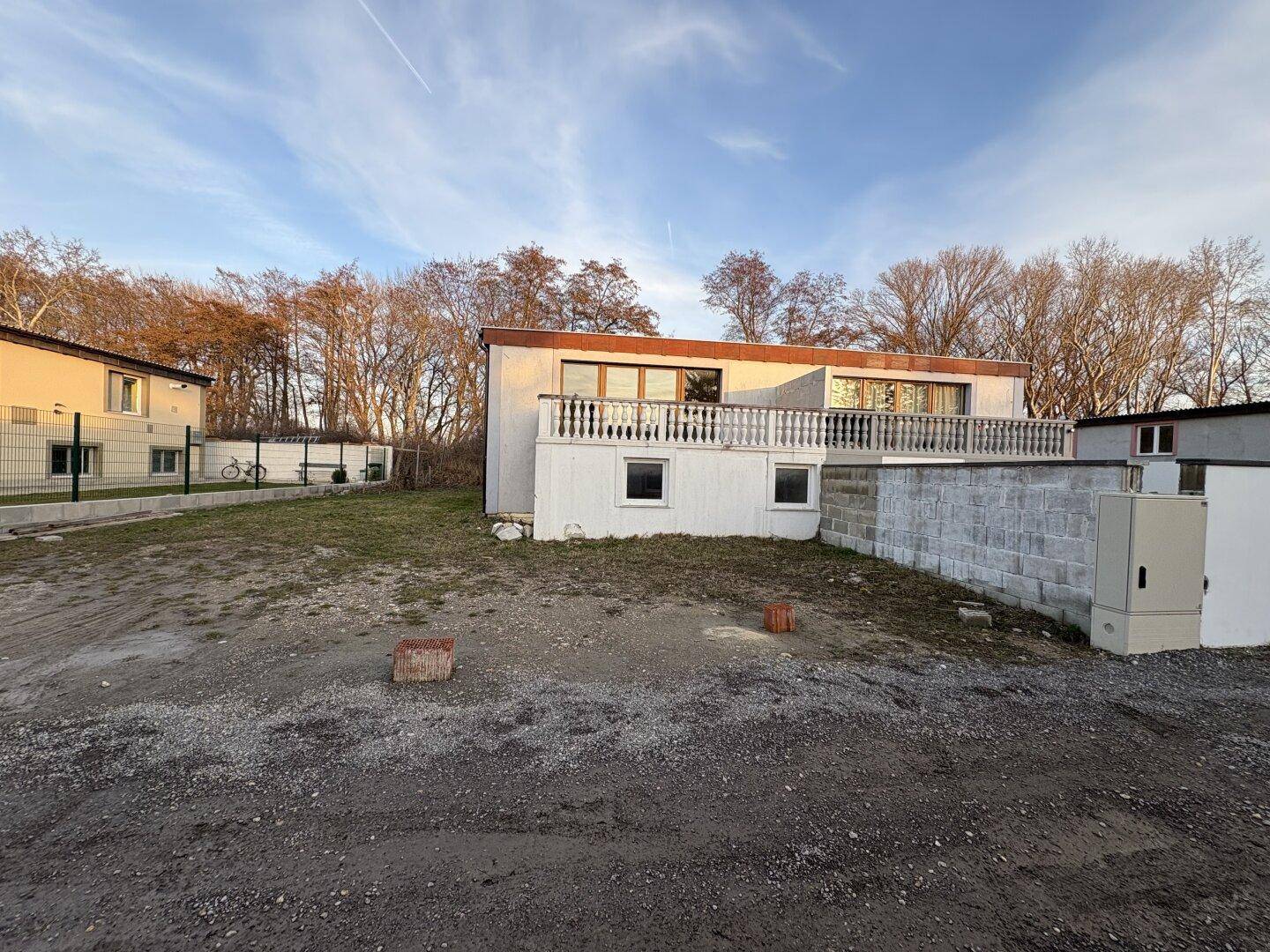 Seitenansicht des Hauses mit Balkon und Balustrade, umgeben von unbefestigtem Grundstück und Bäumen.