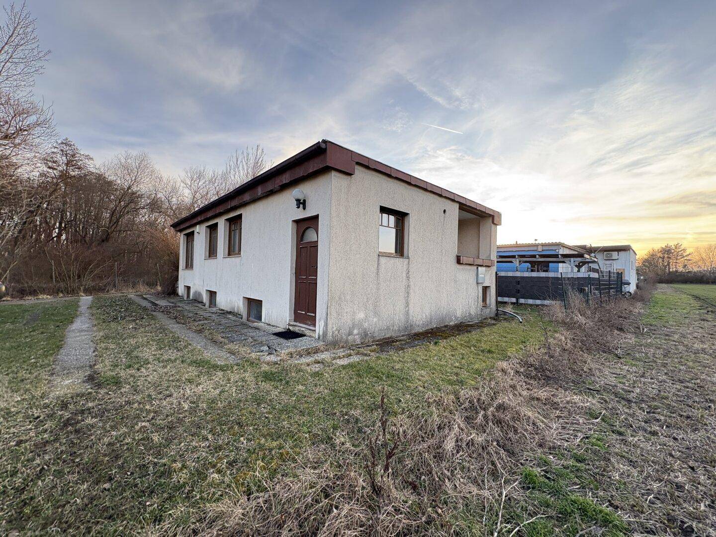Einfaches, älteres Haus mit Putzfassade und braunen Fenstern in ländlicher Umgebung.