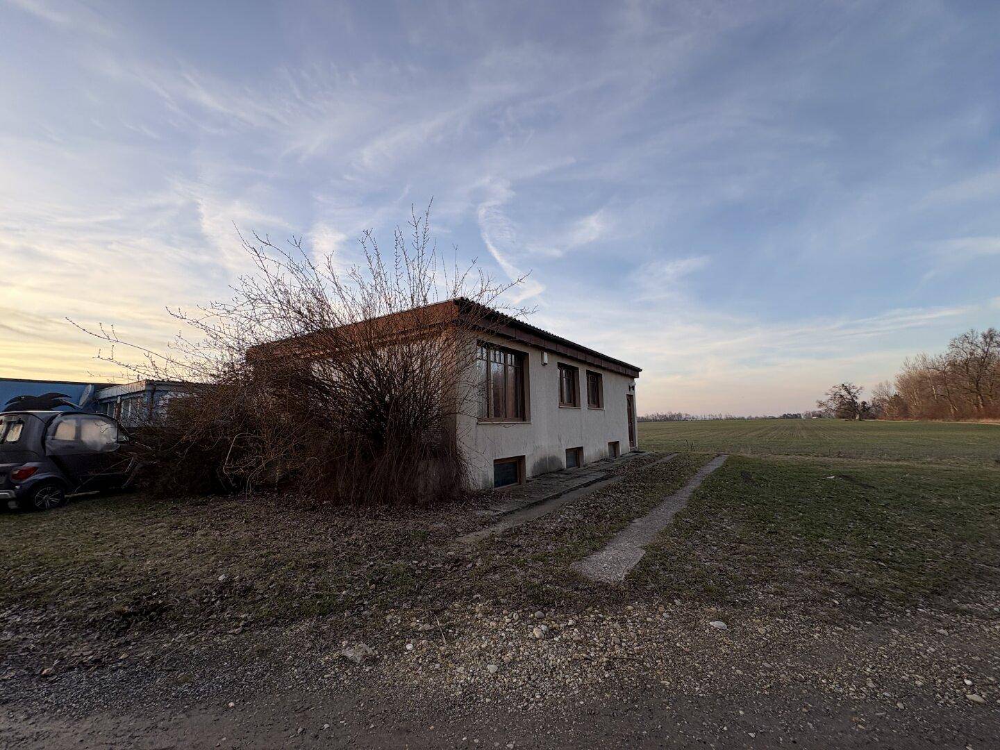 Das Haus von der Seite, umgeben von einer weiten Wiese unter einem bewölkten Himmel.