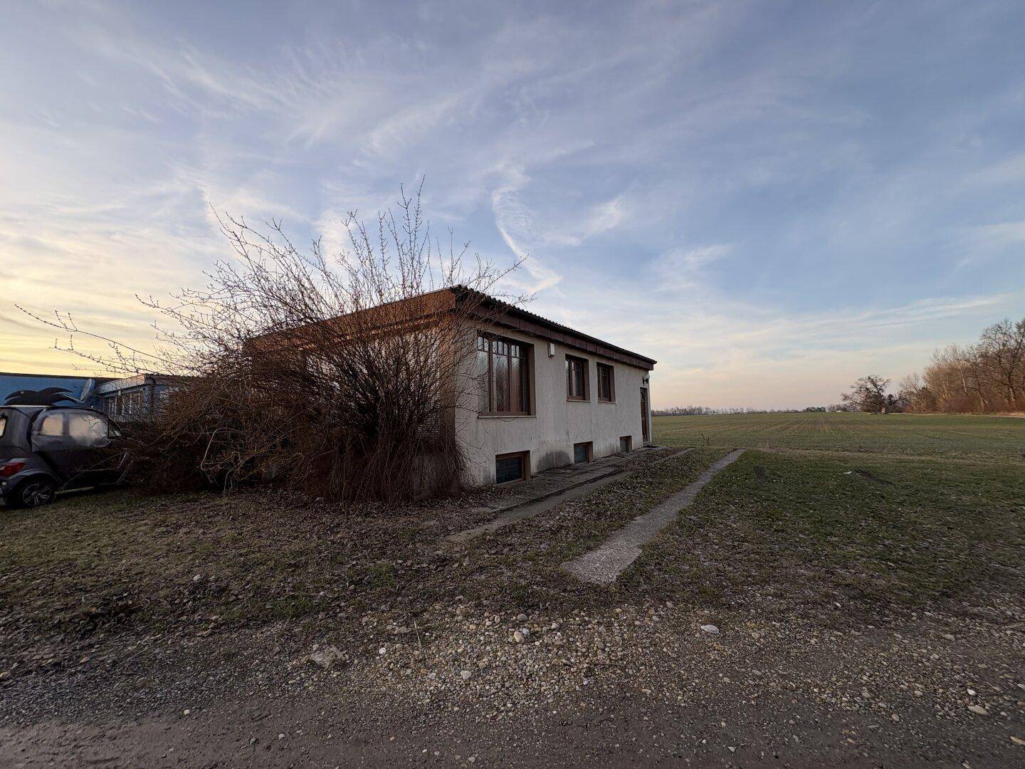 Das Haus von der Seite, umgeben von einer weiten Wiese unter einem bewölkten Himmel.