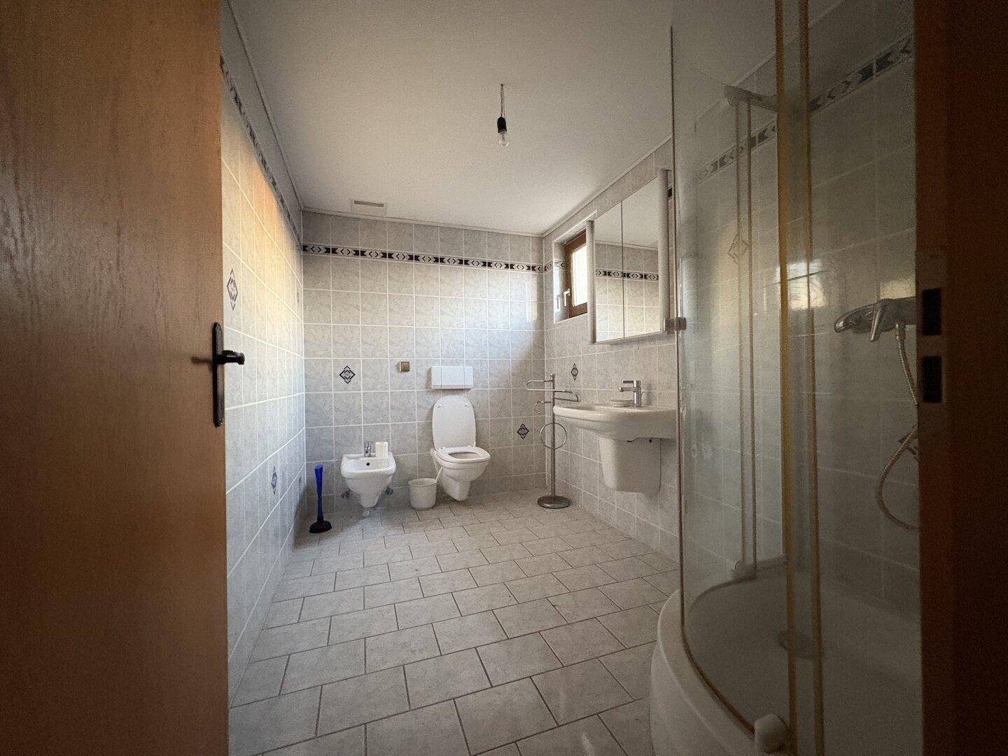 Gefliestes Badezimmer mit Dusche, WC, Bidet und Waschbecken in hellem Design.