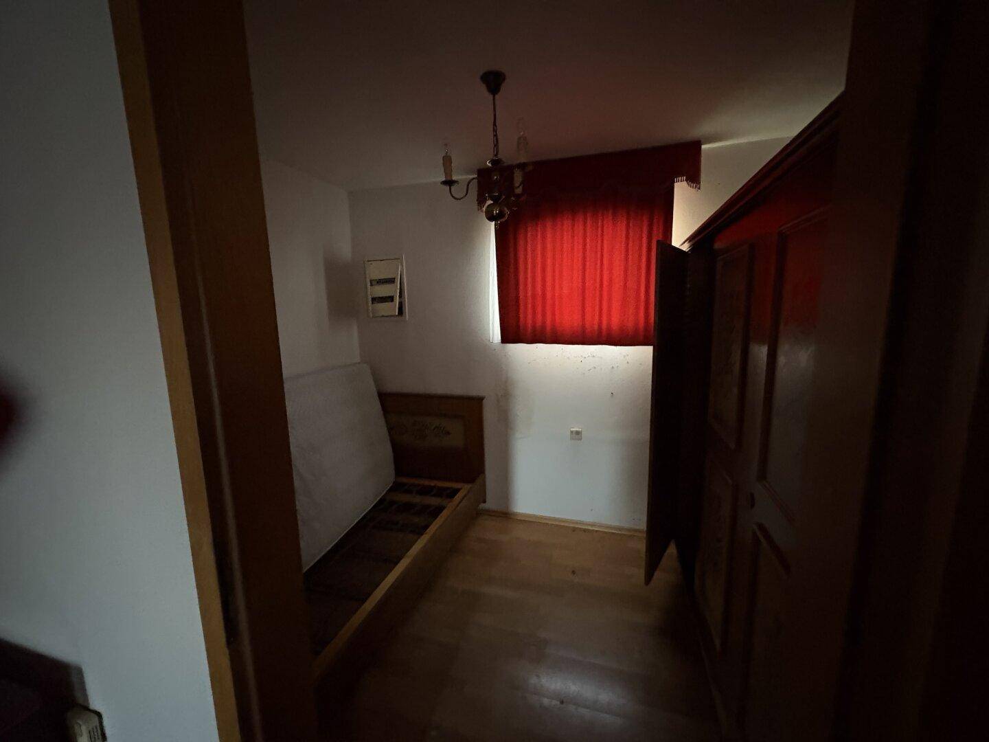 Kleines Schlafzimmer mit Holzbettrahmen, dunklem Kleiderschrank und rotem Vorhang.