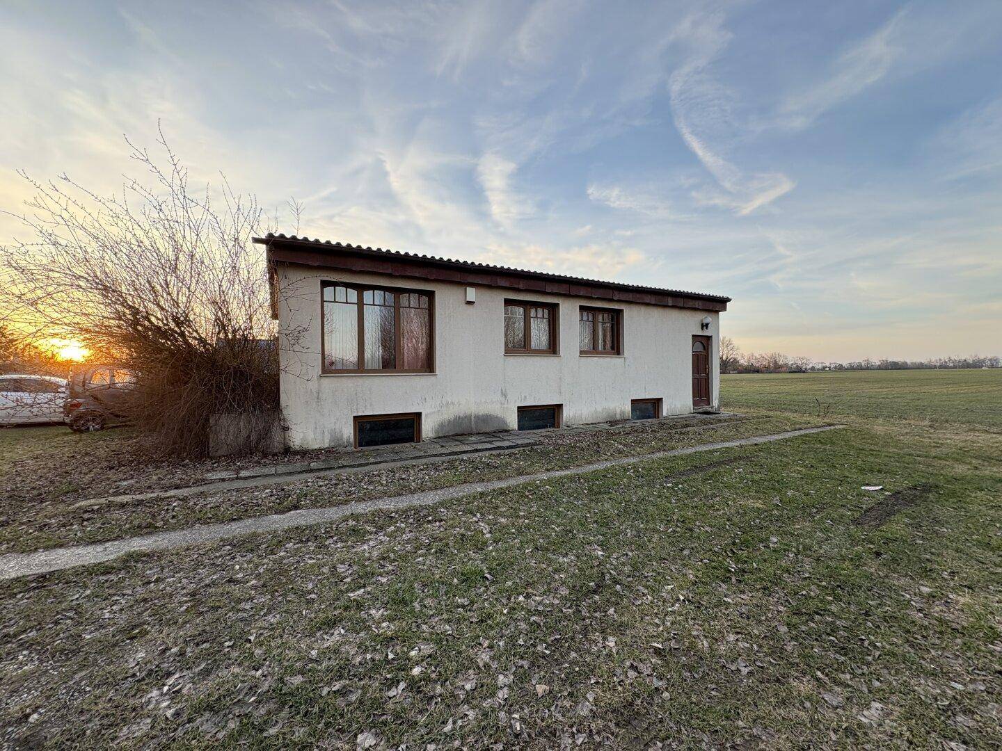Seitenansicht des Hauses mit einfacher Architektur und Blick auf die angrenzende Feldlandschaft.
