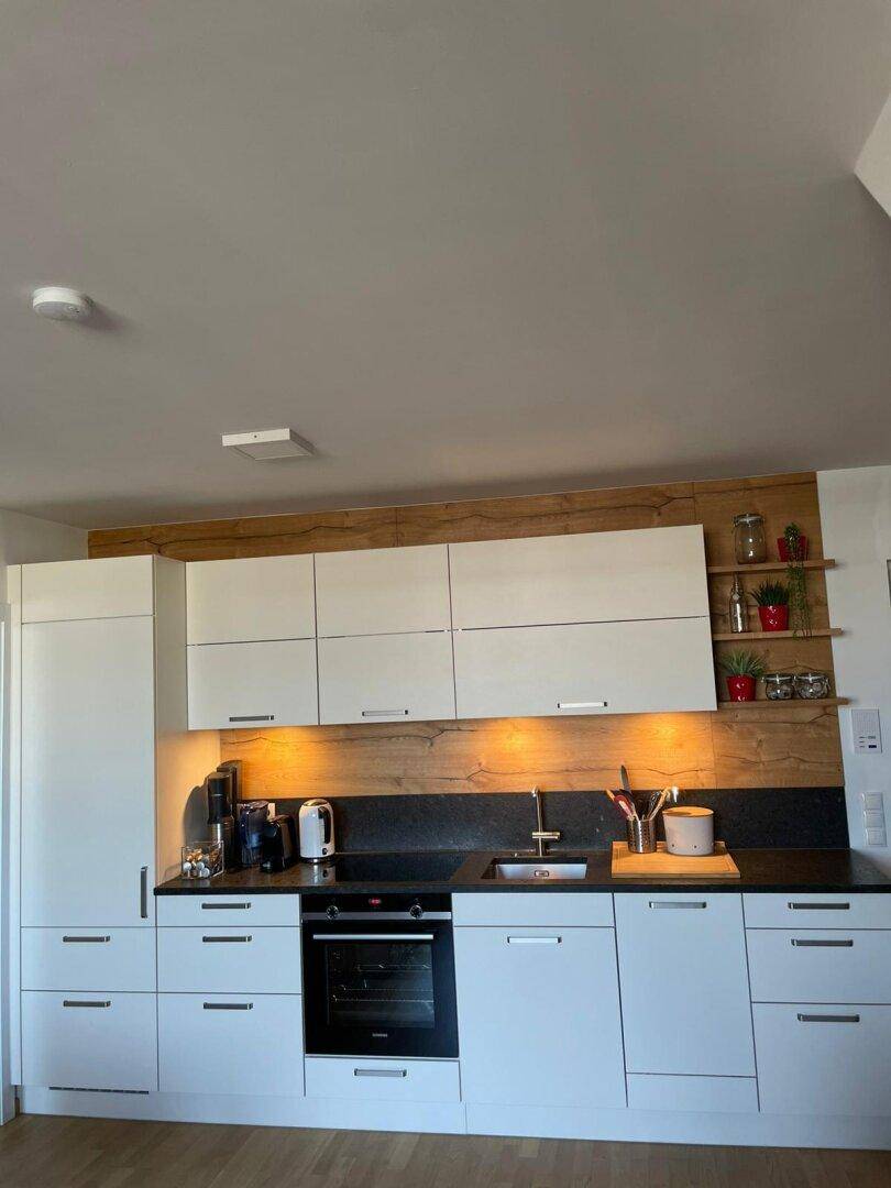 Moderne Einbauküche mit weißen Schränken, Holzdetails und dunkler Arbeitsplatte, gut ausgestattet.