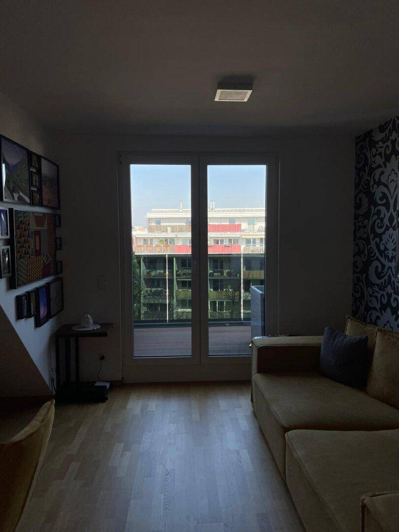 Wohnzimmer mit Sofa, Holzboden und großem Fenster mit Blick ins Grüne und Zugang zur Terrasse.