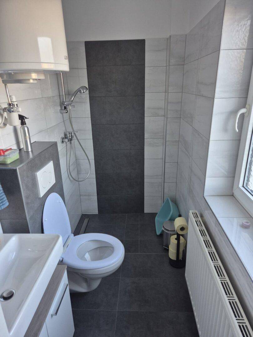 Modern gefliestes Badezimmer mit Toilette, Waschbecken, Duschbereich und Fenster für Tageslicht.