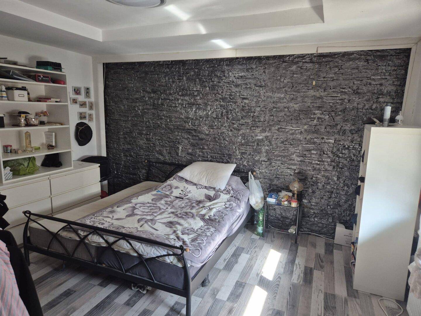 Schlafzimmer mit Einzelbett, dunkler Strukturwand und Holzoptik-Boden, einfach eingerichtet.