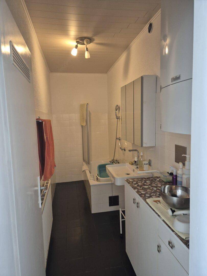 Langes Badezimmer mit Badewanne, Waschbecken, Spiegelschrank und Holzdecke, funktional eingerichtet.