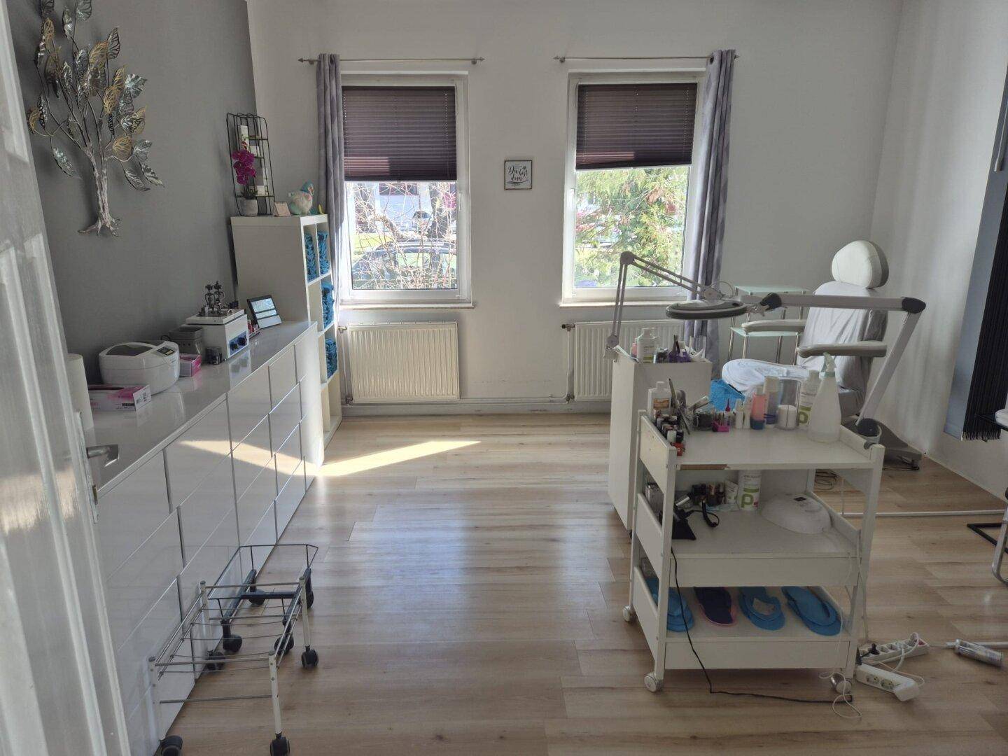 Heller Raum mit Laminatboden, zwei Fenstern und Ausstattung für eine Behandlung oder Büroarbeit.