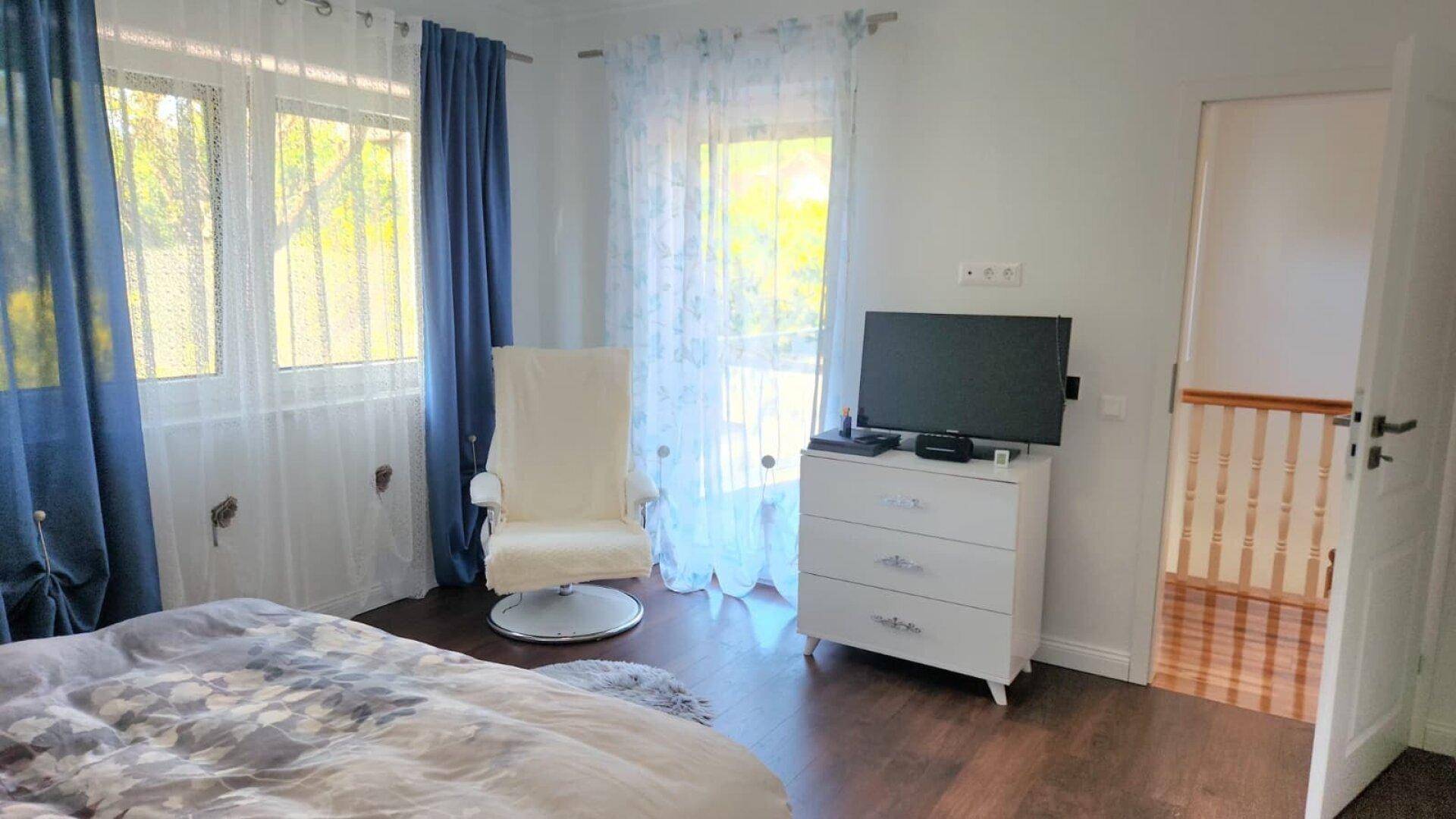 Gemütliches Schlafzimmer mit Bett, Kommode und Fenstern mit blauen Vorhängen.