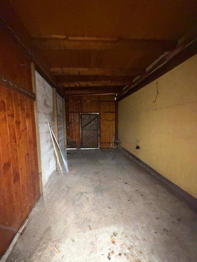 Langer, schmaler Garagen- oder Abstellraum mit Holzwänden und Betonboden, vielseitig nutzbar.