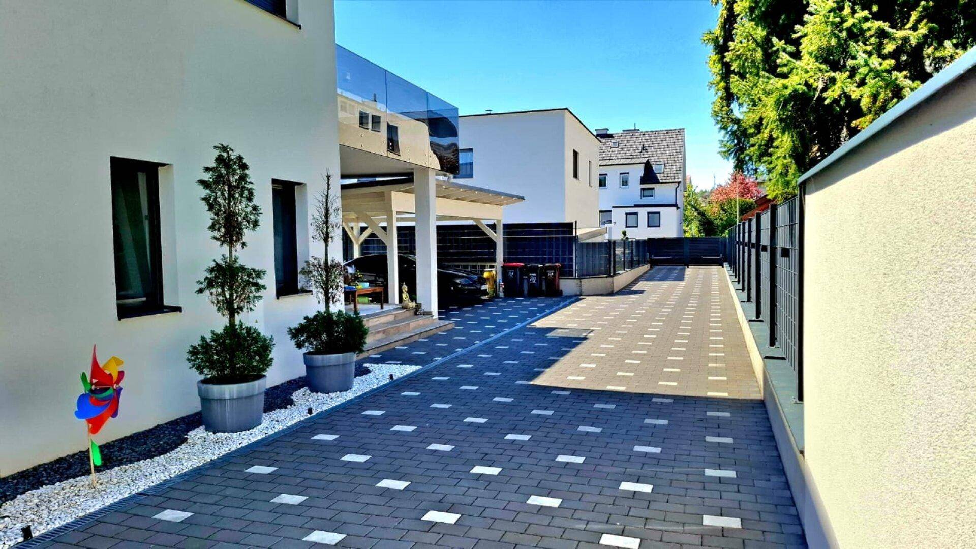 Lange gepflasterte Auffahrt mit modernem Carport und gepflegten Pflanzen entlang des Weges.