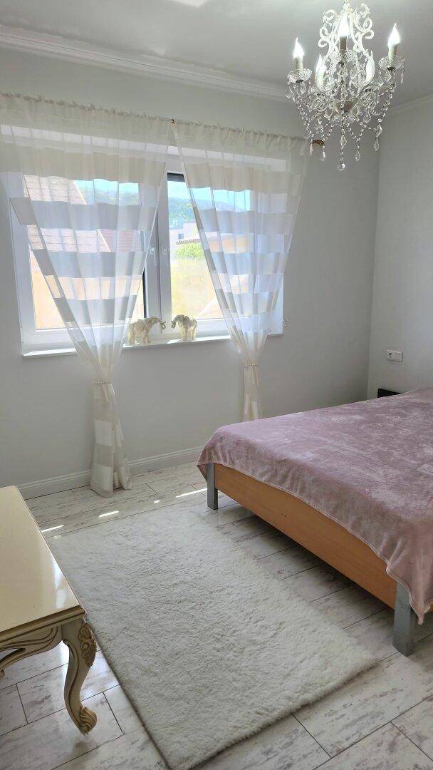 Helles Schlafzimmer mit Doppelbett, hellem Boden und Fenster mit transparenten Vorhängen.