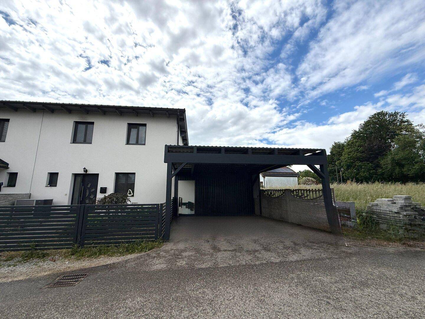 Weißes Reihenhaus mit modernem Carport und angrenzender Grünfläche unter bewölktem Himmel.