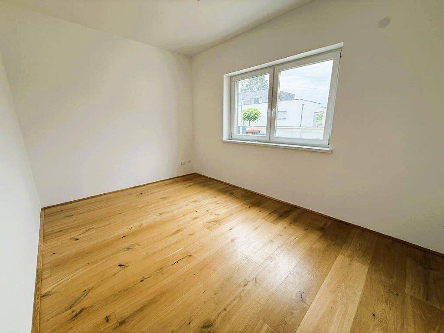 Helles, leeres Zimmer mit Holzboden und einem großen Fenster, vielseitig nutzbar.