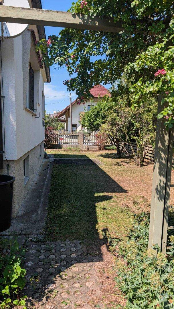 Ein gepflasterter Gartenweg führt entlang der Hausseite zu einem hinteren Gartenbereich mit Zaun.