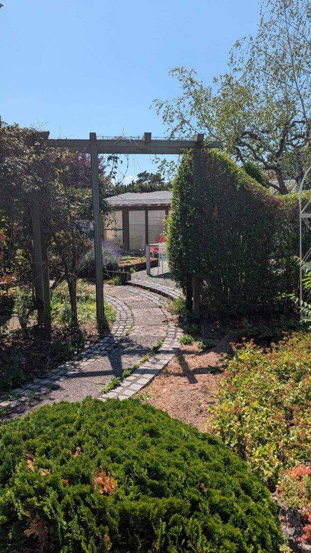 Ein geschwungener Gartenweg führt durch üppiges Grün zu einem Wintergarten im Hintergrund.