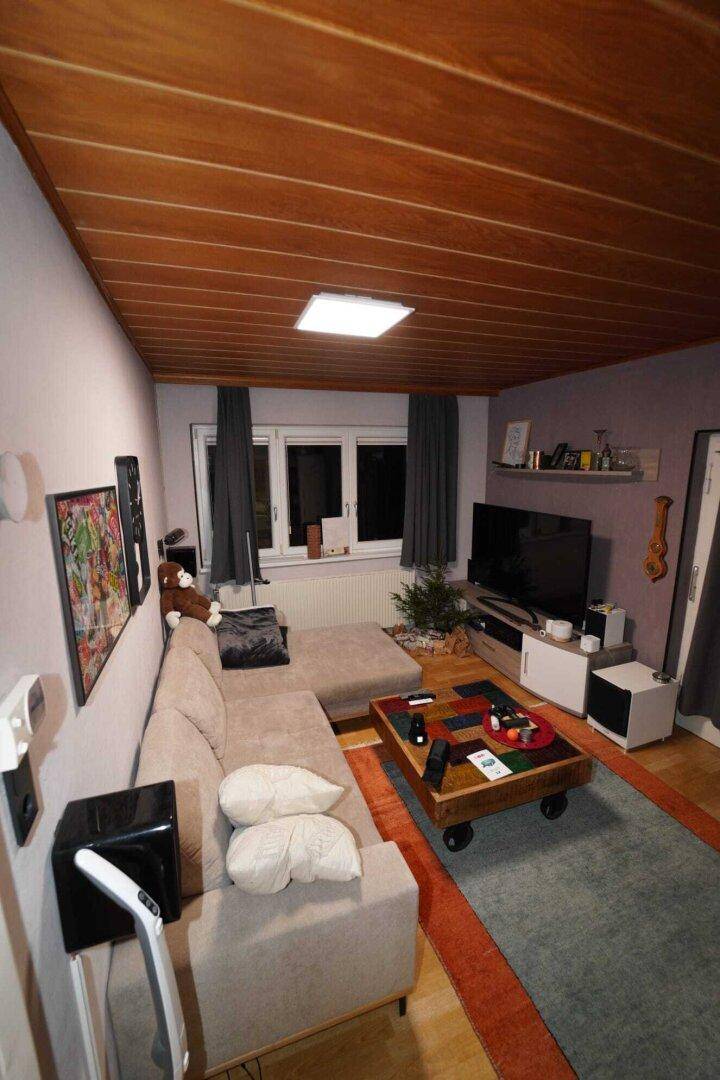 Wohnzimmer mit beigem Sofa, Holzdecke, Parkettboden und Blick nach draußen.