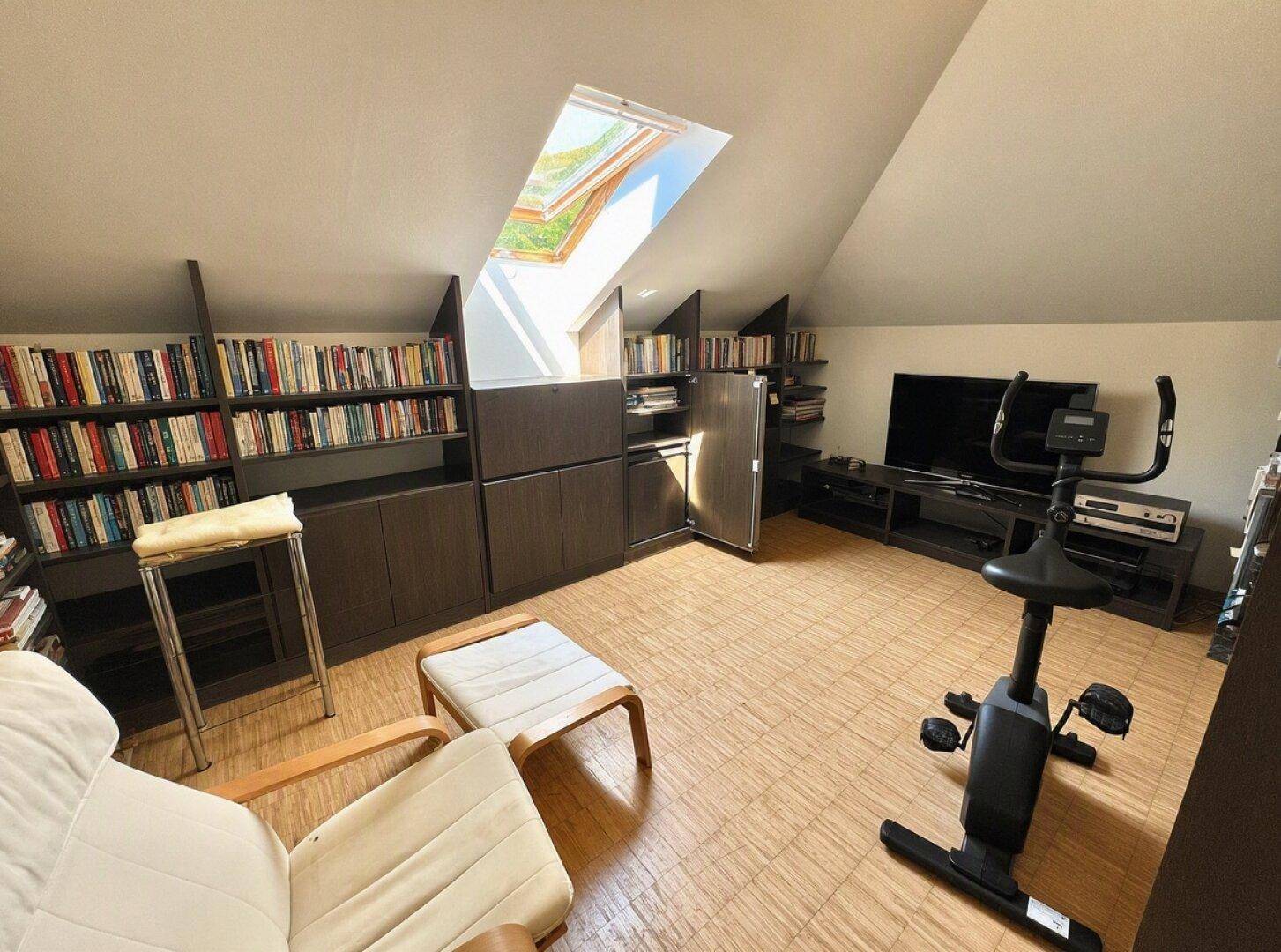 Dachgeschossraum mit Dachschrägen, Bücherregalen und einem Heimtrainer, ideal als Büro.