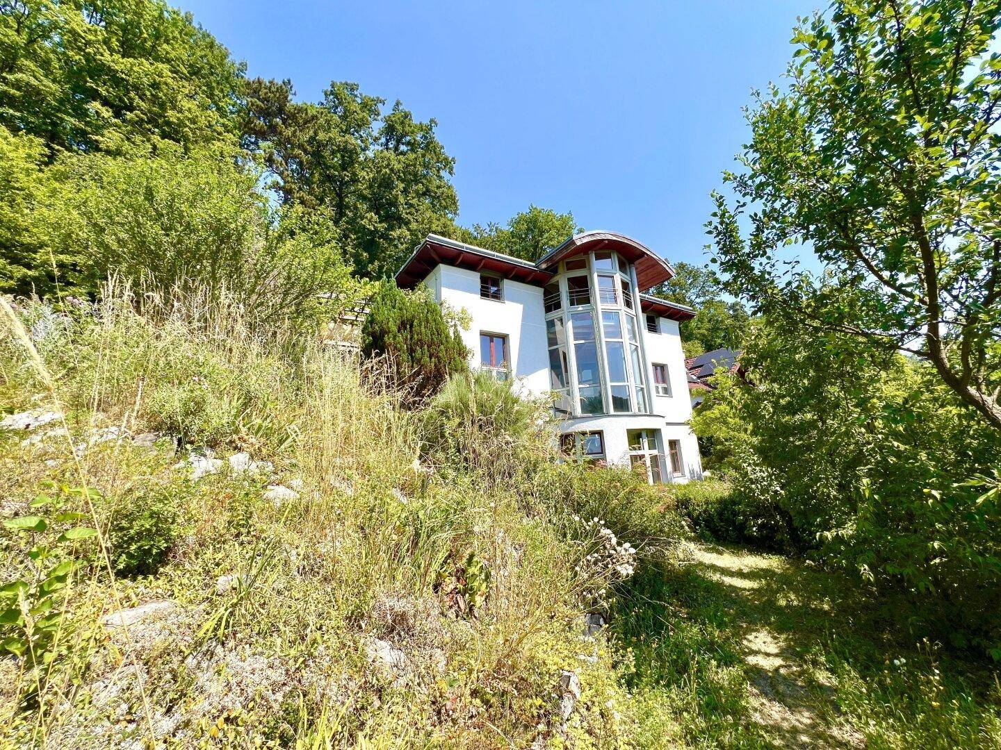 Modernes Wohnhaus eingebettet in eine hügelige, grüne Landschaft mit viel Baumbestand.