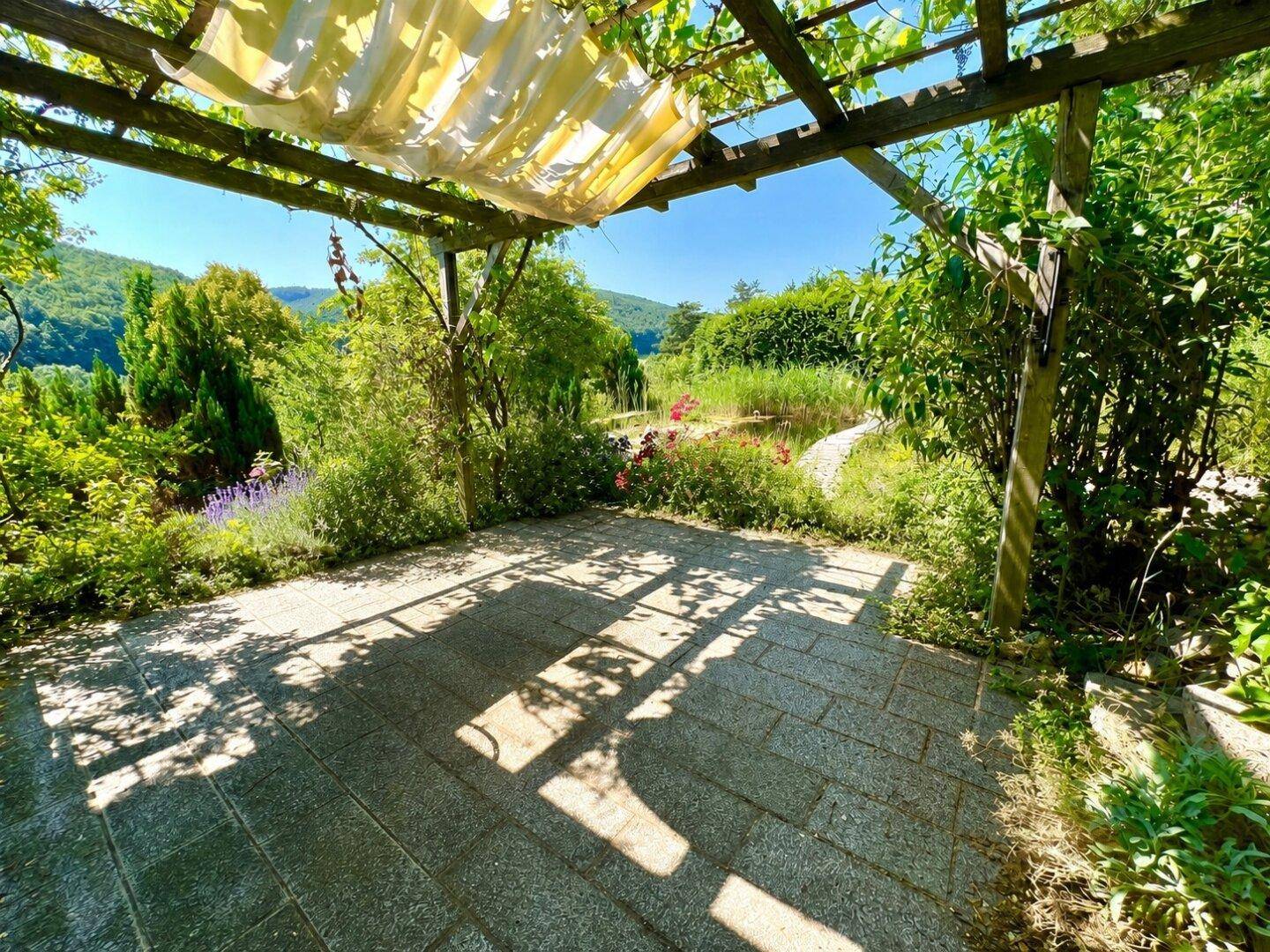 Gemütliche Steinterrasse unter einer Pergola mit Blick auf den Teich und die grüne Landschaft.