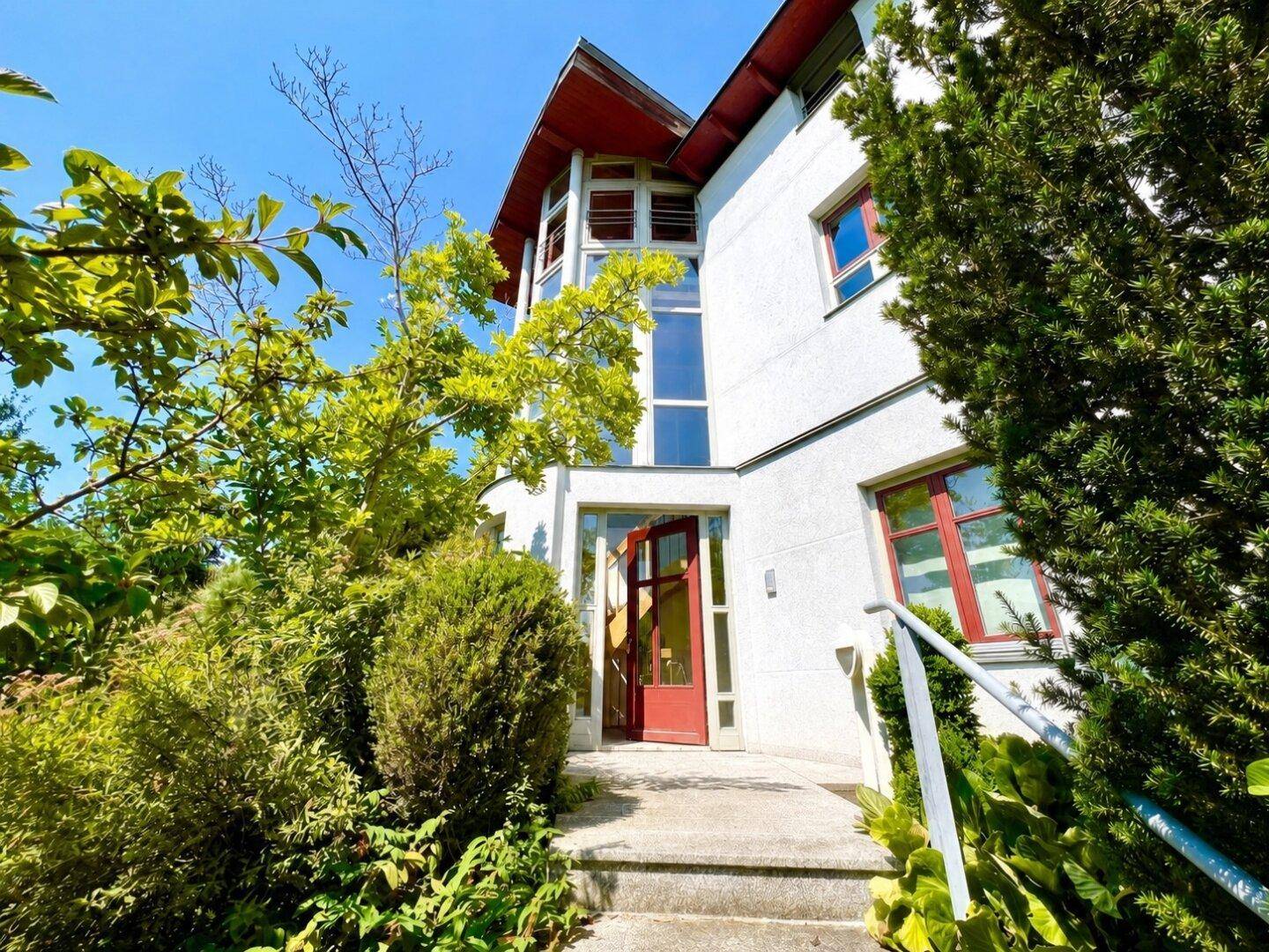 Eingangsbereich des modernen Hauses mit roter Tür und umgebender grüner Bepflanzung.