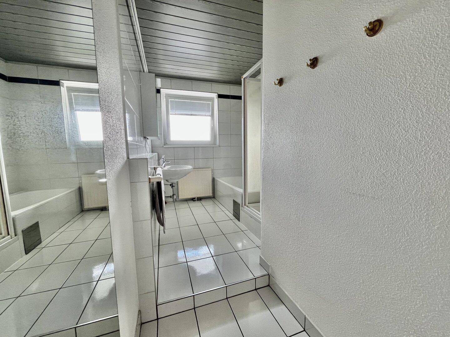 Badezimmer mit weißen Fliesen, Badewanne mit Duschwand, Waschbecken und Fenster.