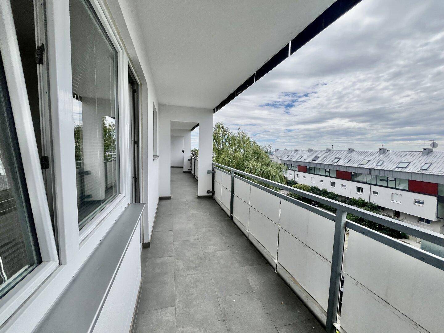 Langer Balkon mit Fliesenboden und Blick auf die umliegenden Gebäude und Bäume.