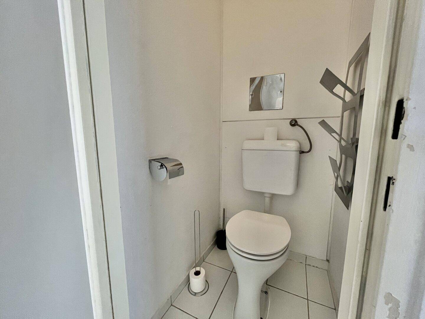 Separates WC mit Toilette, kleinem Spiegel und weißen Fliesen an Boden und Wänden.