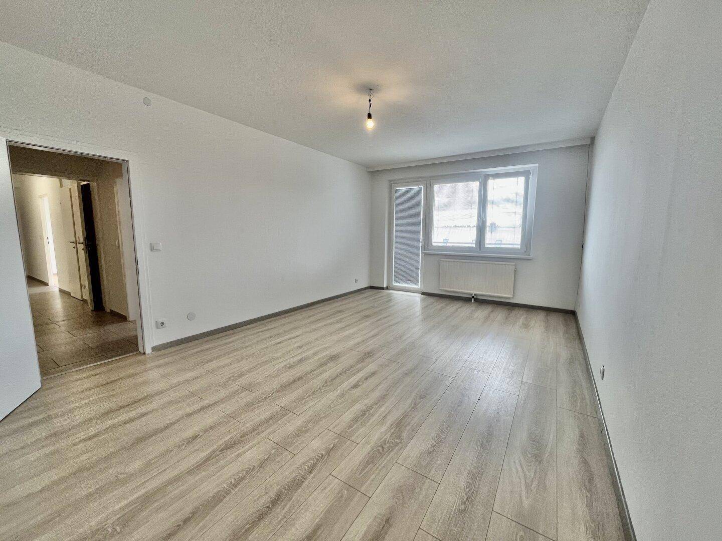 Geräumiges Wohnzimmer mit hellem Laminatboden, großen Fenstern und Zugang zum Balkon.