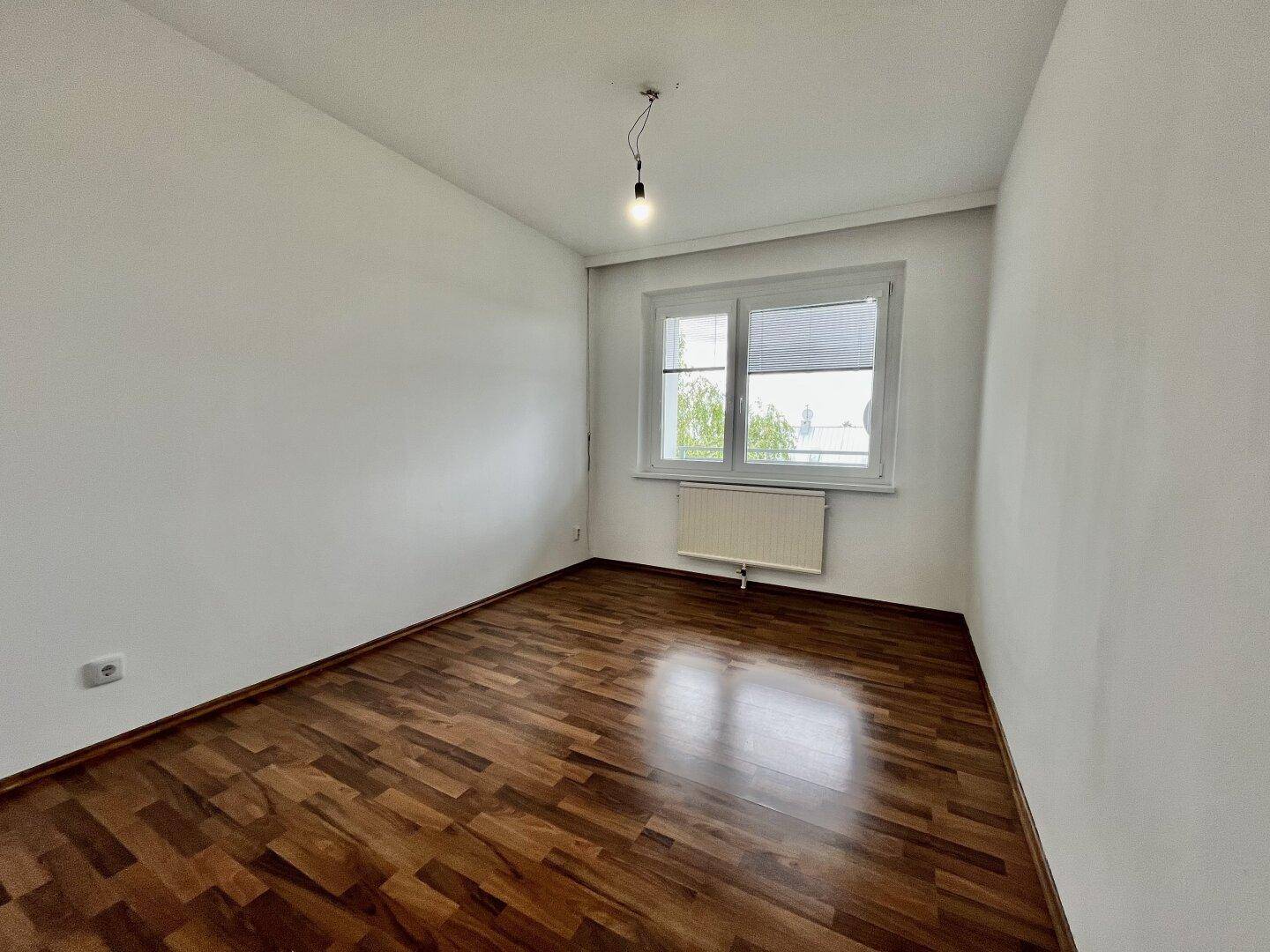 Gemütliches Zimmer mit dunklem Parkettboden, weißen Wänden und einem Fenster für Tageslicht.