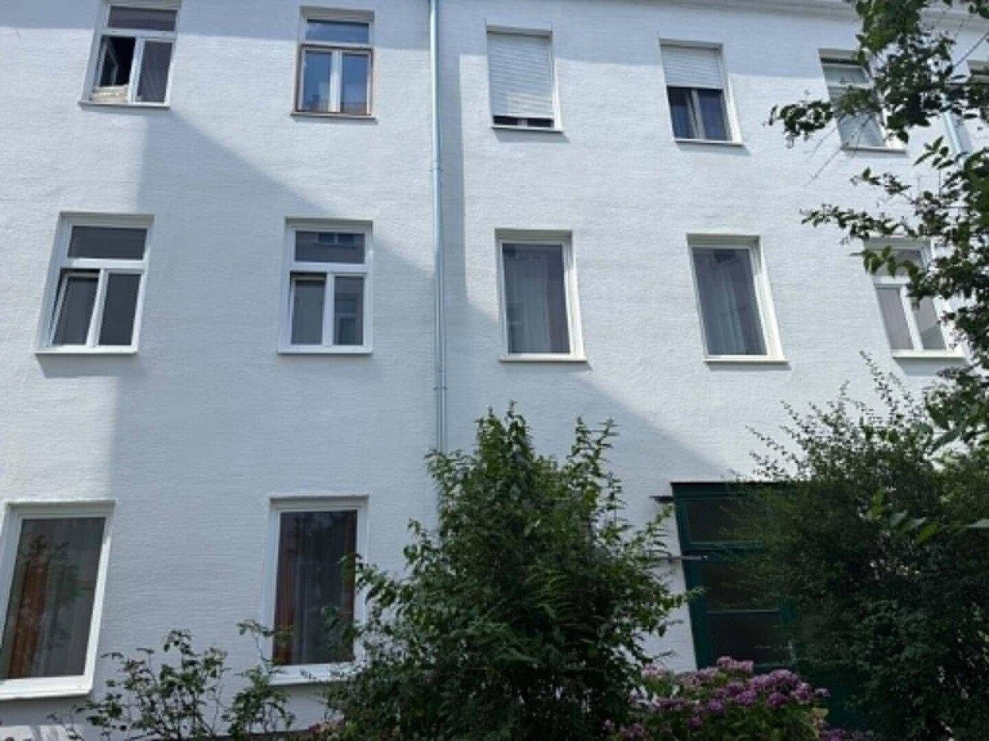 Weiße Gebäudefassade mit mehreren Fenstern und grünen Büschen im Vordergrund.