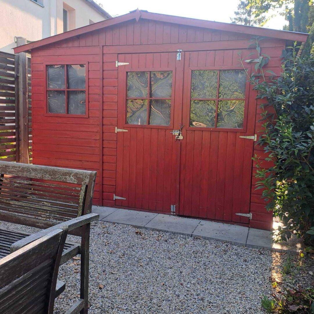 Roter Holzschuppen im Garten mit Doppeltür und kleinen Fenstern neben einer Sitzbank.