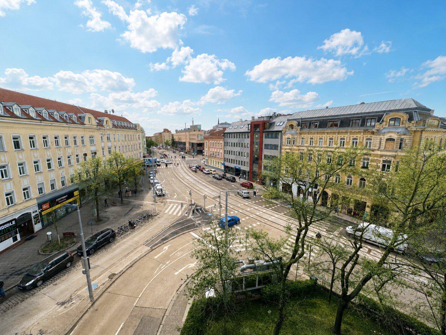Panoramablick auf eine belebte Stadtstraße mit historischen Gebäuden, Straßenbahnschienen und grünen Bäumen unter blauem Himmel.