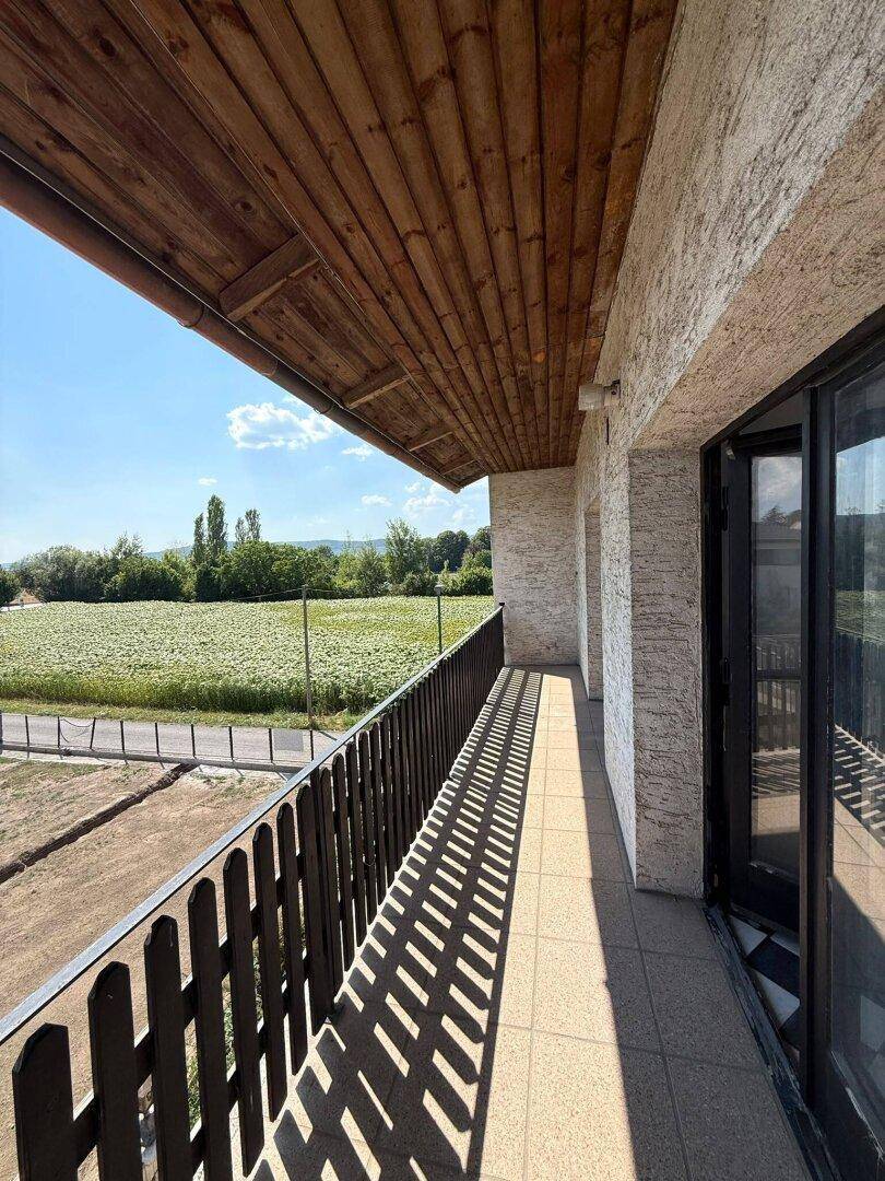 Langer Balkon mit Holzdach und Blick auf weite Felder unter strahlendem Sonnenschein.