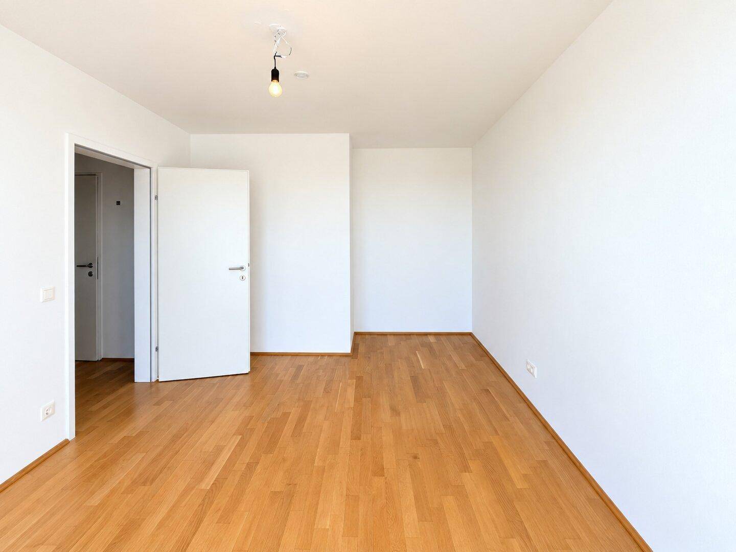 Leeres, helles Zimmer mit Parkettboden und weiß gestrichenen Wänden.