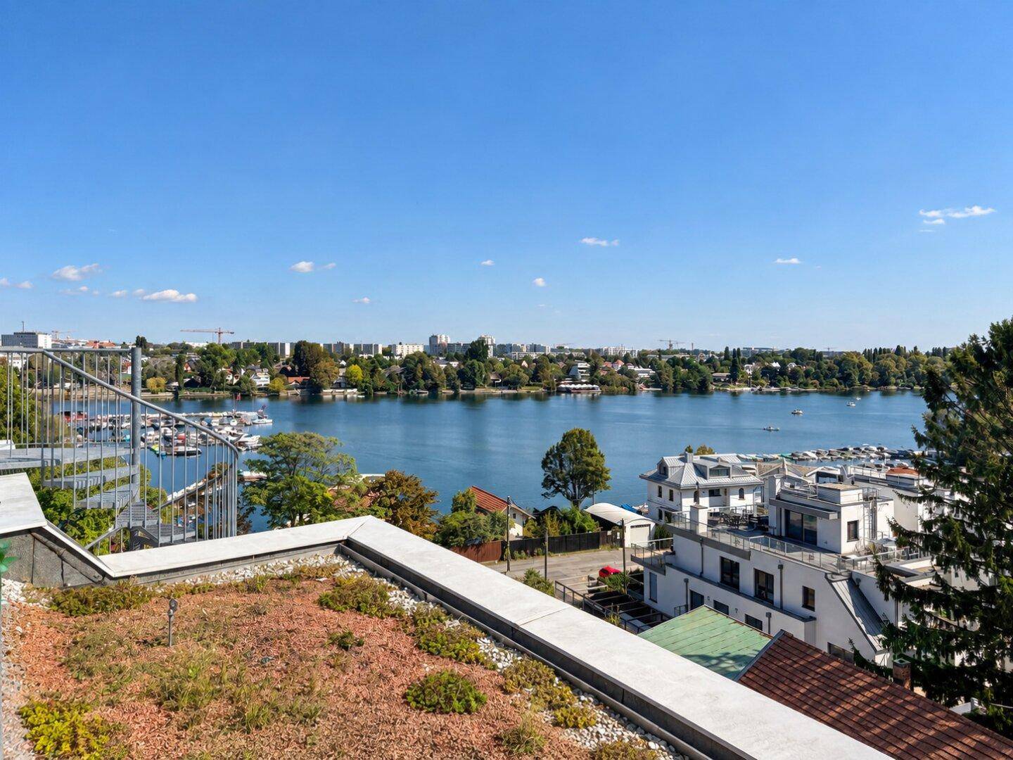 Panoramablick über einen See mit Yachthafen und begrüntem Dachbereich im Vordergrund unter blauem Himmel.