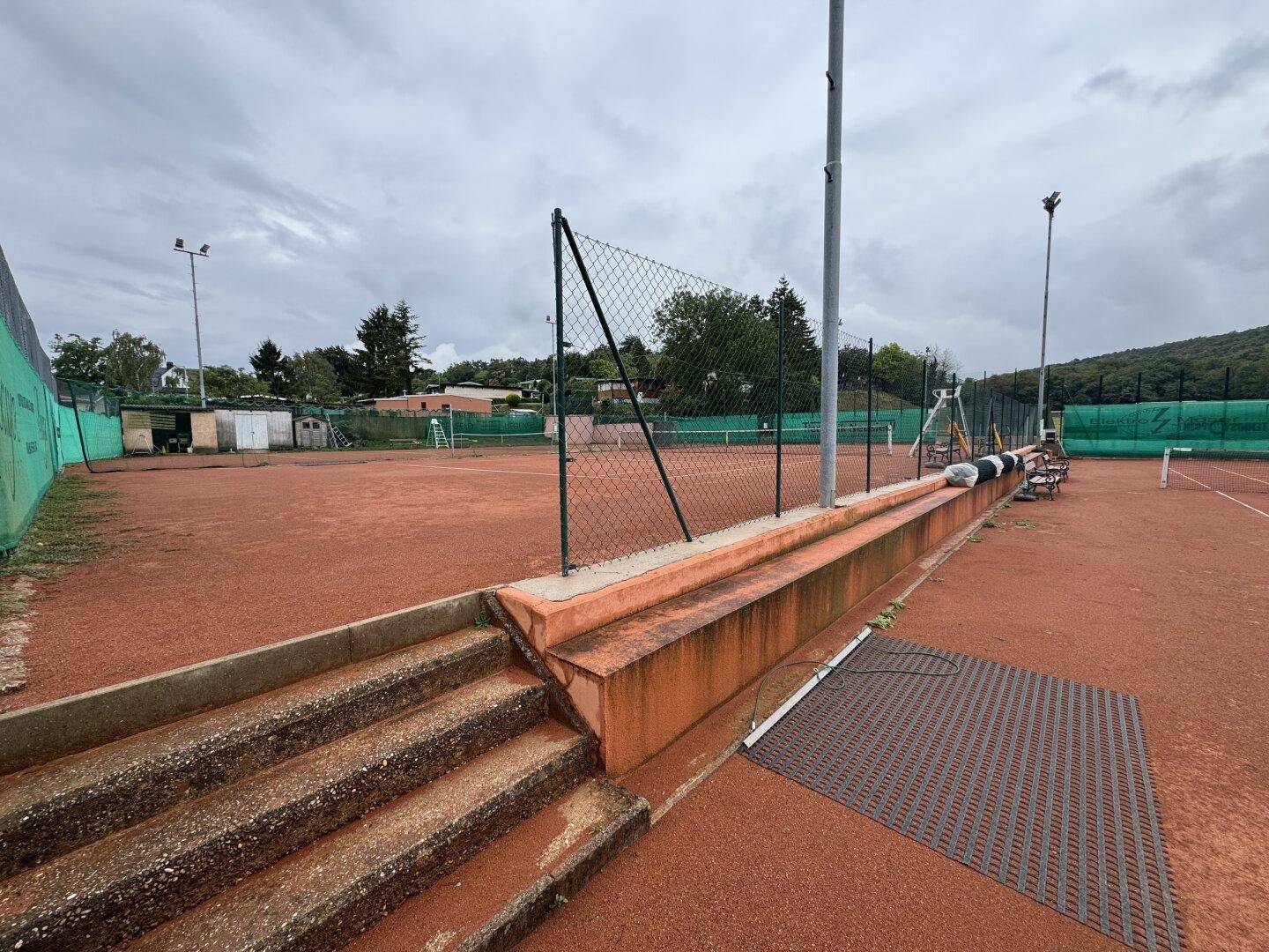 Tennisplätze mit rotem Sandbelag und Zugangstreppe, umgeben von grüner Landschaft.