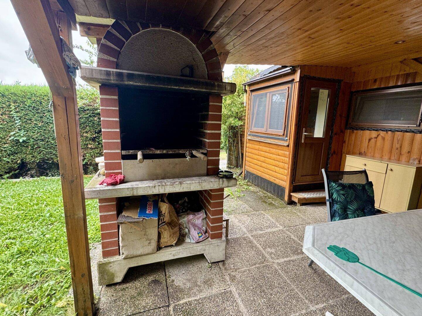 Überdachte Terrasse mit gemauertem Grill und Fliesenboden, ideal für Mahlzeiten im Freien.