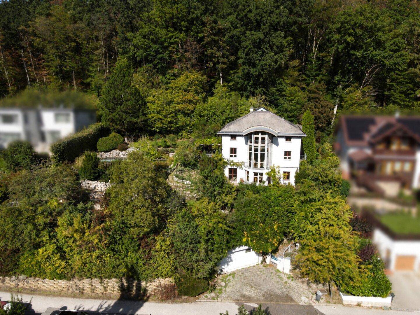 Weitläufige Luftaufnahme des Hauses, eingebettet in eine grüne Hügellandschaft mit Wald im Hintergrund.