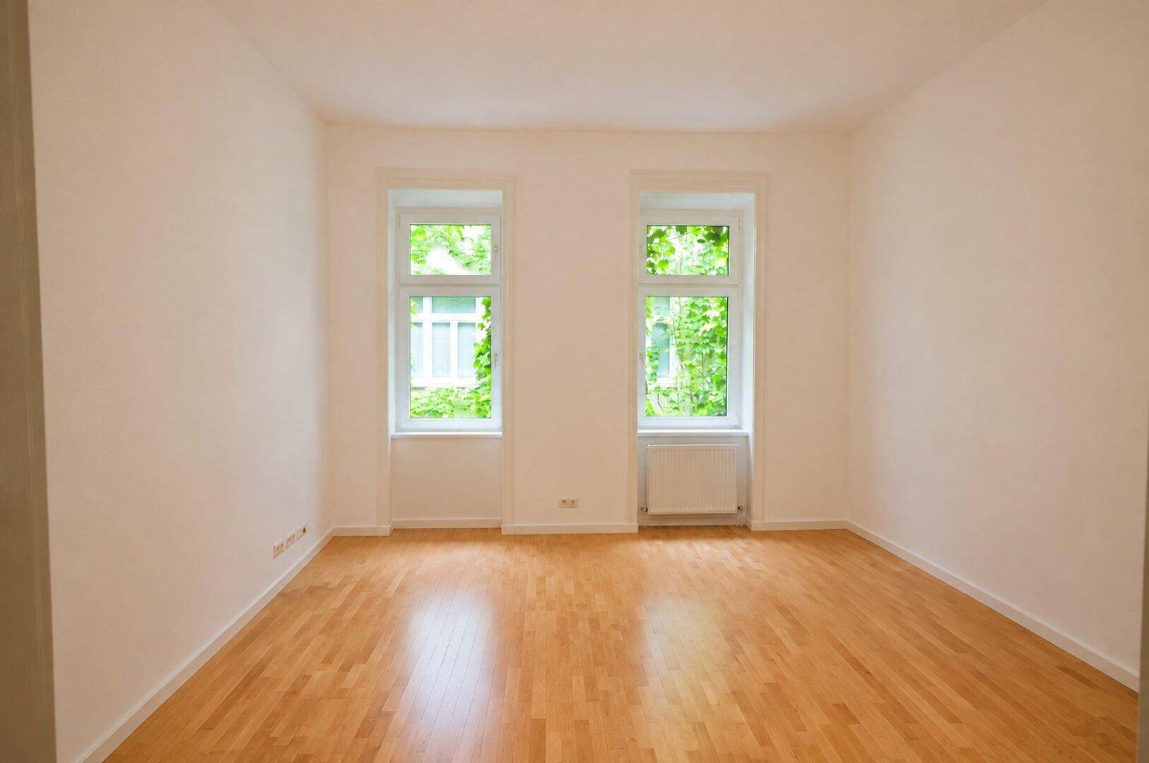 Geräumiges, helles Zimmer mit zwei Fenstern, Holzboden und weiß gestrichenen Wänden.