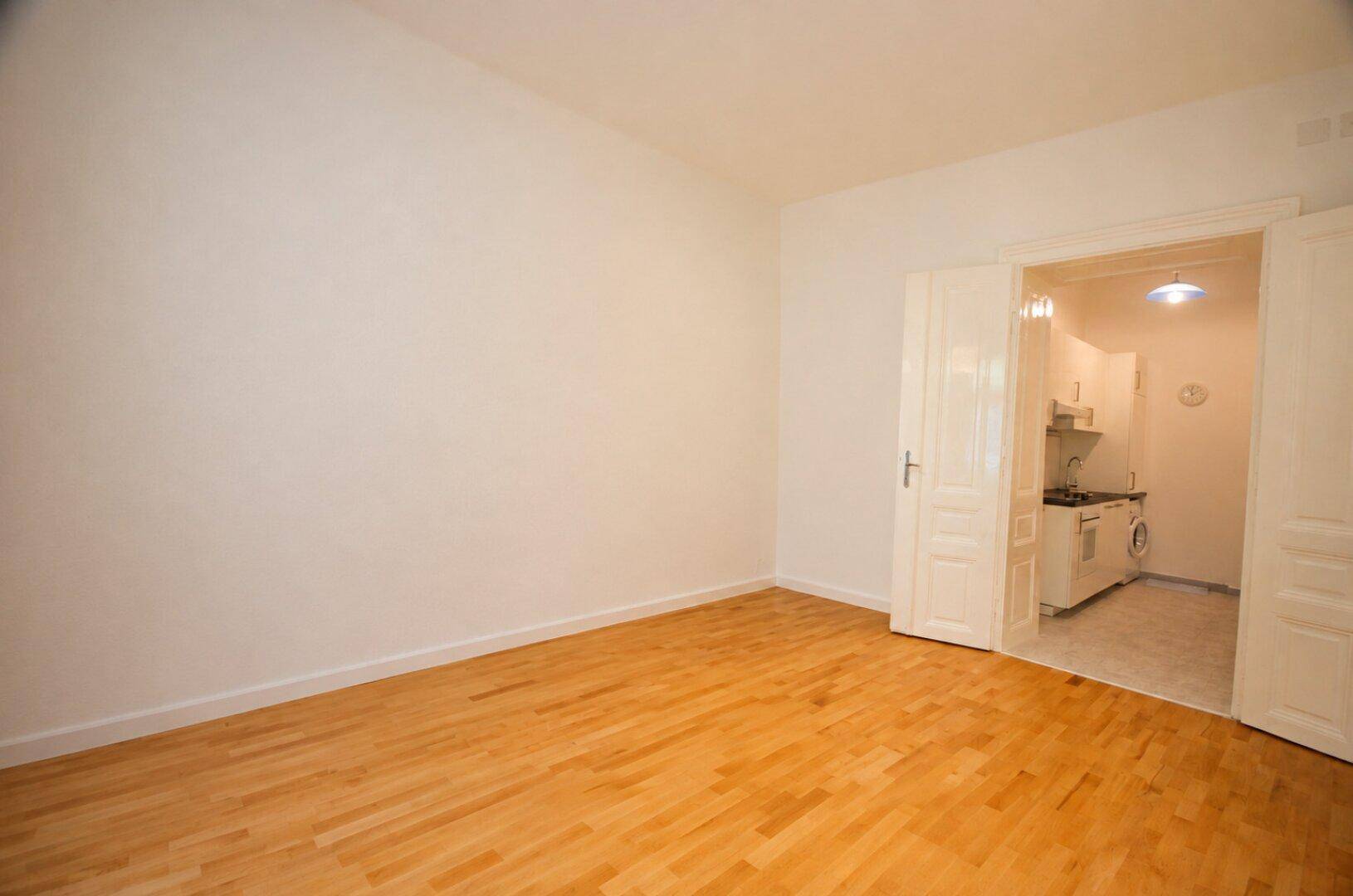 Helles Zimmer mit Holzboden und Doppeltür, die zu einer kleinen Küche führt.