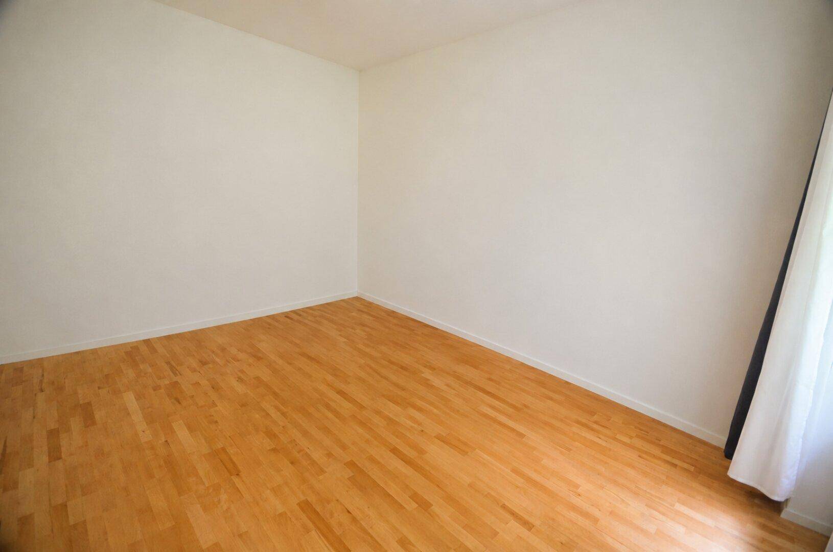 Leeres Zimmer mit Holzboden, weißen Wänden und einem Fenster mit Vorhang.
